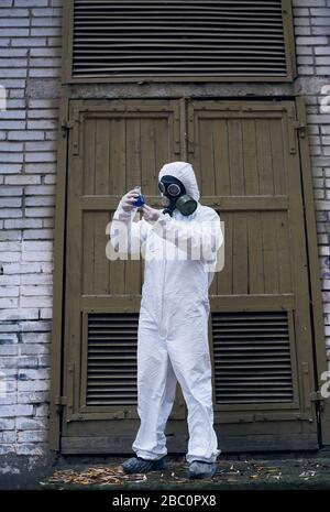 Full length snapshot of ecologist standing in front of old door and observing reaction in glass flask with blue liquid, laboratorial experiment. Scientist wearing white uniform, gloves and gas mask Stock Photo