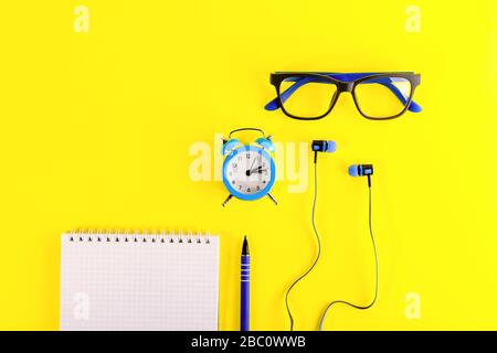 Small blue alarm clock, in minimalist style, blue earphones, blue Eyeglasses and note book with blue pen, lies  on bright yellow background. Remote wo Stock Photo
