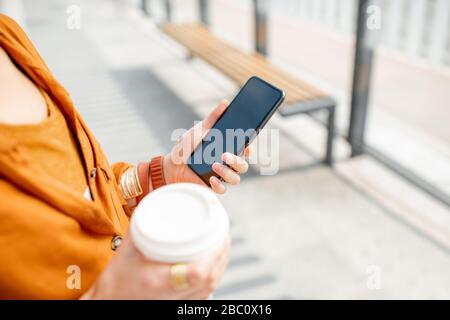 Business woman holding smart phone and coffee cup outdoors, close-up on hands, phone with black screen Stock Photo