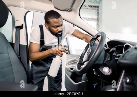 Young African male worker in uniform cleaning car interior and steering wheel with special cleaning chemical spray. Chemical and wet cleaning, car Stock Photo