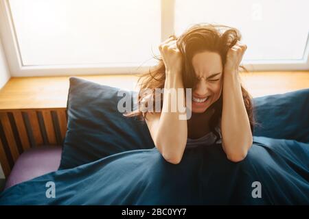 Young beautiful woman in morning bed at home. Hold hands on head close to ears and suffer from pain and headache. Strees and painful morning. Stock Photo