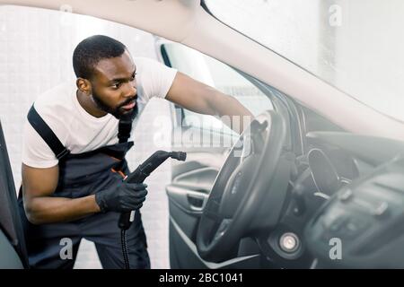 Cleaning of car salon with steam cleaner. Side view of professional male African worker cleaning car interior, steering wheel and control panel using Stock Photo