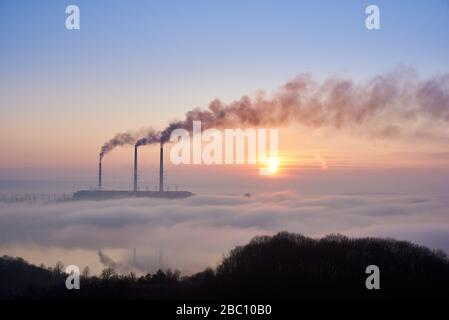 Horizontal snapshot of three smoking stacks of thermal power station on the horizon taken from the hill, pipes are in evening fog on blue sky, copy space. Concept of environmental pollution Stock Photo