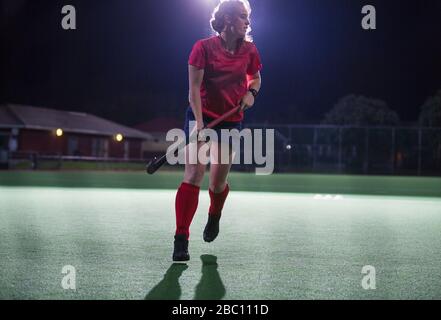 Young female field hockey player running with hockey stick on field at night Stock Photo