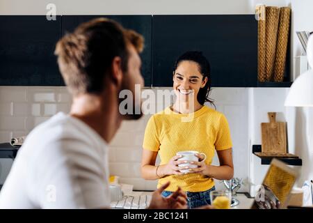 Portrait of young woman smiling at boyfriend with laptop in the kitchen Stock Photo