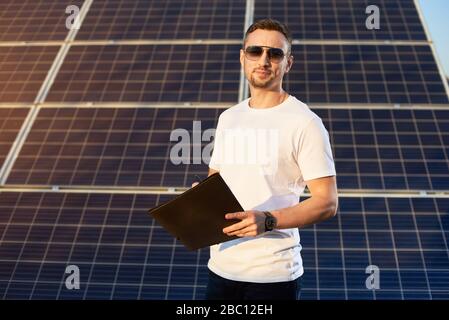 Attractive man with a folder in his hands on a background of solar panels on a warm day. A young man in sunglasses in a white T-shirt and black pants is looking at the camera with a smile Stock Photo