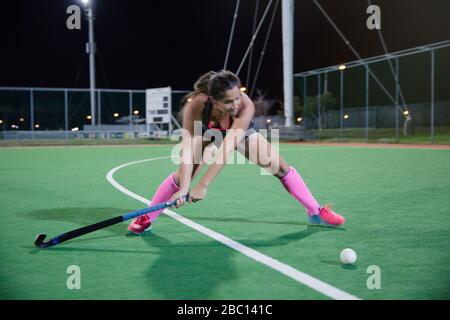 Determined young female field hockey player hitting the ball on field at night Stock Photo