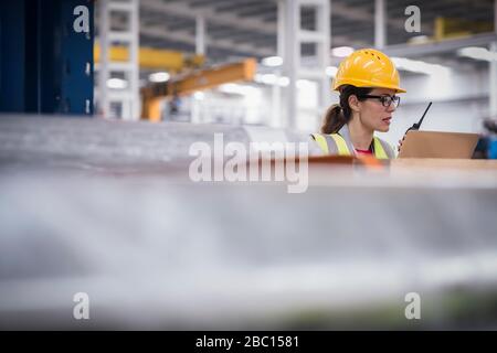 Female worker using laptop and walkie-talkie in factory Stock Photo