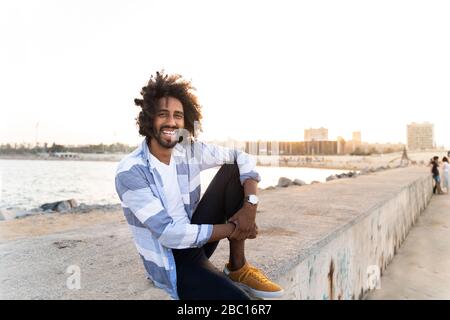 Portrait of happy young man sitting on quay wall at sunset