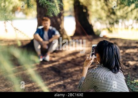 Young woman taking cell phone picture of boyfriend under a tree Stock Photo