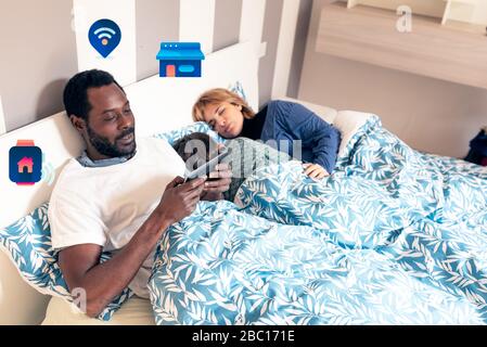 multiethnic family in bed just woke up in the morning, mother and daughter still sleeping, black dad uses the tablet for remote control of the house, Stock Photo