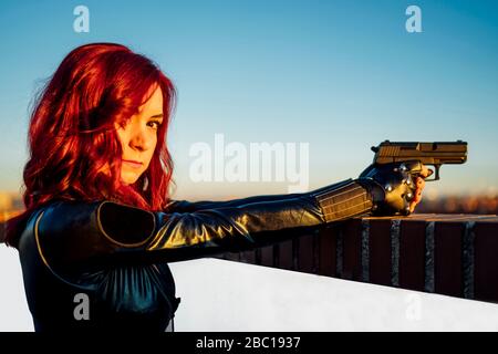 Portrait of redheaded woman with gun in her hand wearing black leather catsuit on rooftop Stock Photo