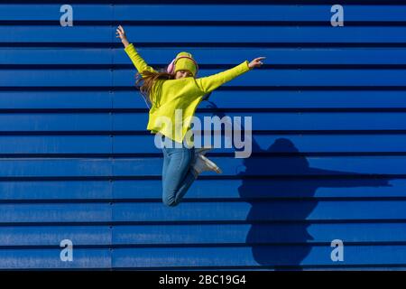 Teenage girl jumping in the air in front of blue background listening music with headphones Stock Photo