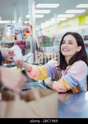 Smiling, friendly female cashier giving receipt to customer at supermarket checkout Stock Photo