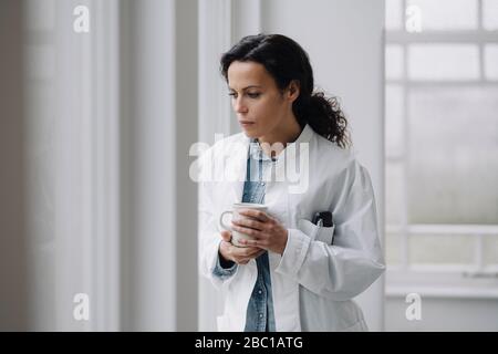 Female doctor standing at the window, looking worried, holding cup of coffee Stock Photo