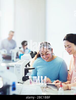 Smiling female science teacher and girl student reading textbook at microscope in laboratory classroom Stock Photo