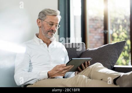 Senior man with grey hair in modern design living room sitting on couch using tablet Stock Photo
