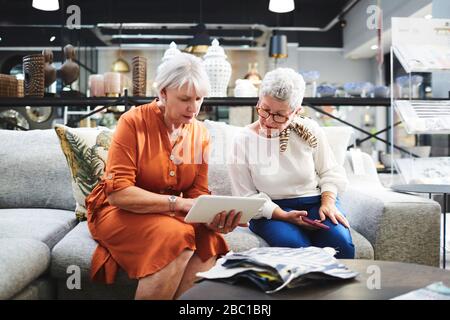 Senior women with digital tablet looking at fabric swatches on sofa in furniture store Stock Photo