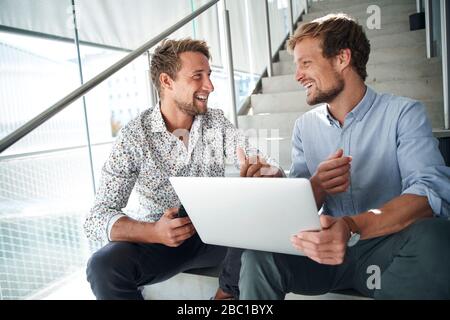Two happy young businessmen sitting on stairs using laptop Stock Photo