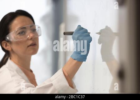 Female science teacher wearing lab coat, rubber glove and goggles, writing at whiteboard in classroom Stock Photo