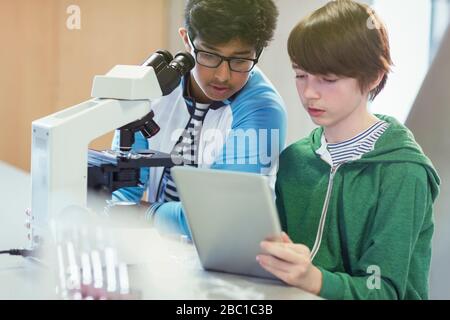 Focused boy students using digital tablet at microscope in laboratory classroom Stock Photo