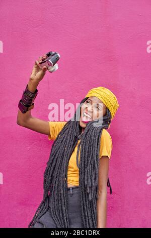 Portrait of woman with long dreadlocks taking a selfie with her camera in front of a pink wall Stock Photo