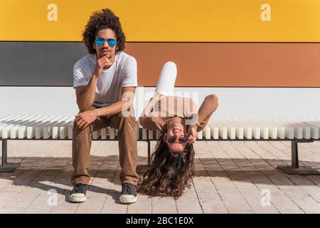 Couple with sunglasses, relaxing on bench, one partner lying on back with head hanging down Stock Photo