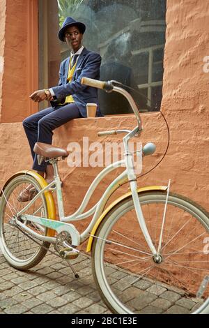 Stylish young businessman with bicycle wearing old-fashioned suit sitting on a the ledge of a window Stock Photo