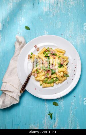 Italian recipe Pasta tortilloni with green pea, mint leaves, cheece, smoked bacon and cheese. Top view, blue background. Stock Photo