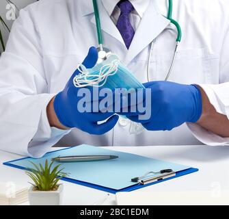doctor in a white coat and blue latex gloves sits at a white desk and shows a stack of disposable medical masks, protective agent against viruses and Stock Photo