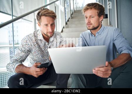 Two young businessmen sitting on stairs using laptop Stock Photo