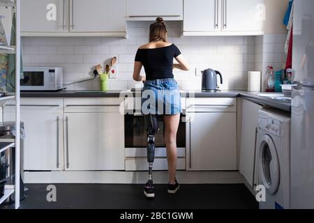 Rear view of young woman with leg prosthesis in kitchen at home Stock Photo