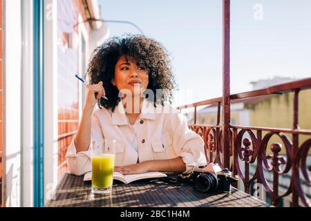 Smiling young woman with curly hair sitting at table on balcony with notebook, smoothie and camera Stock Photo
