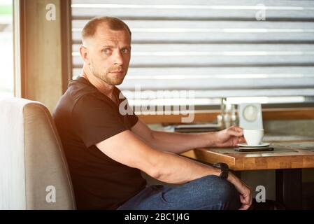 Handsome thoughtful sporty man is holding a cup of coffee, sitting at the window side table in cafe, wearing black t-shirt and dark blue jeans Stock Photo