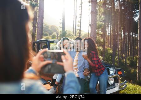 Young woman with camera phone photographing friends at jeep in woods Stock Photo