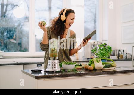 Fit woman standing in kitchen, preparing healthy smoothie, using online recipe