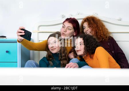 Mother and her three daughters sitting together on bed taking selfie with smartphone Stock Photo