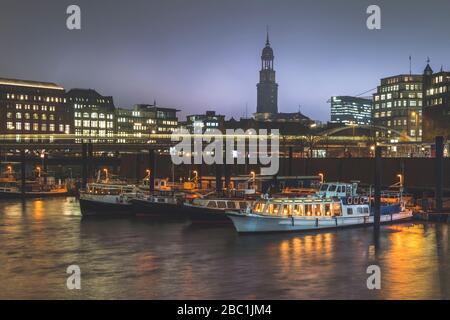 Germany, Hamburg, Boats moored in city harbor at dusk with light trail and tower of Saint Michaels Church in background Stock Photo