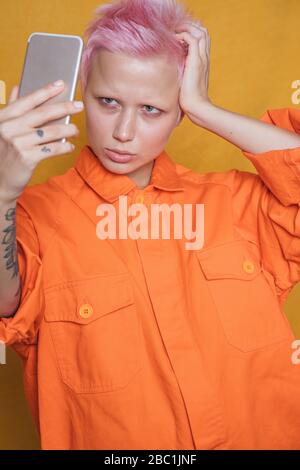 Portrait of young woman with short pink hair wearing orange jacket taking selfie with smartphone Stock Photo