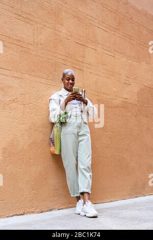 Smiling woman leaning against a wall with thermos flask and groceries using smartphone Stock Photo