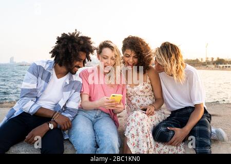 Happy friends sitting on quay wall at sunset using smartphone Stock Photo
