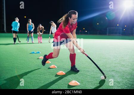 Determined young female field hockey player practicing sports drill on field at night Stock Photo