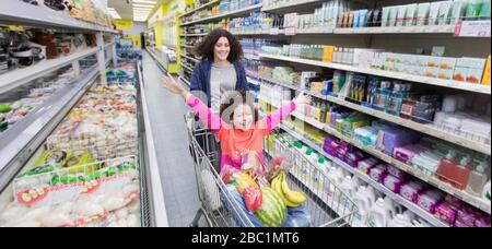 Mother pushing playful daughter in shopping cart in supermarket aisle Stock Photo
