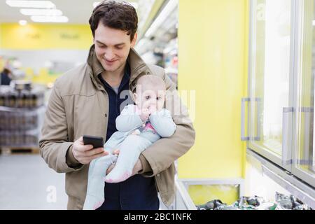 Father with baby daughter checking smart phone, shopping in supermarket Stock Photo