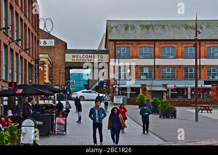 People walking alongside cafés & restaurants with entrance to Gloucester Quays retail shopping centre with more restaurants, behind. Stock Photo