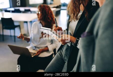 Close-up of a woman making notes while standing in a meeting with colleagues. Female writing on a notepad during a meeting. Stock Photo