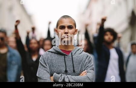 Young woman protestor standing with her arms crossed and group of people in background on the city street. Activists demonstrating in the city. Stock Photo