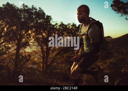 Male runner wearing hydration pack running outdoors over rocky trail on the mountain. Trail runner practicing on mountain path. Stock Photo