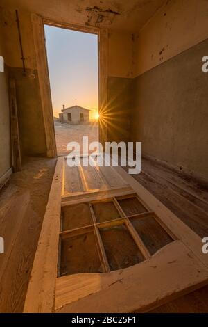 A dramatic vertical photograph inside an abandoned house at sunset, with an old door lying on the floor and a sunburst and house outside, taken in the Stock Photo