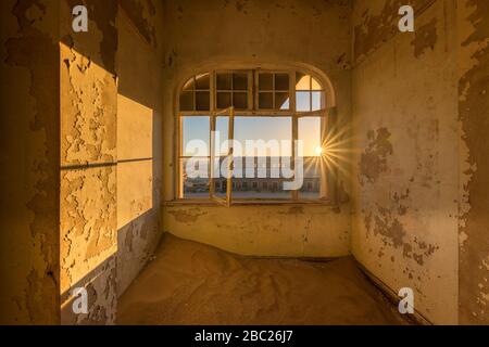 A dramatic photograph inside an abandoned house at sunrise, with a golden sunburst and building through broken windows, taken in the ghost town of Kol Stock Photo